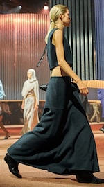 Placeholder for: Model walking on a runway wearing a dark, sleeveless top and long skirt. Others in similar attire are in the background.