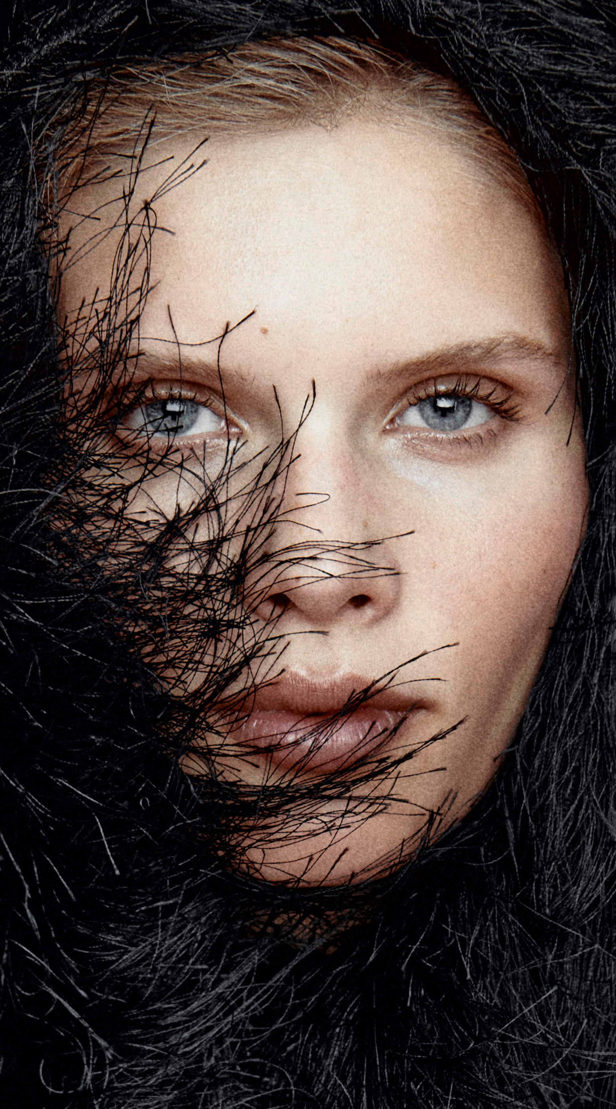 Close-up portrait of a woman's face partially covered by dark, wispy strands, highlighting her blue eyes and intense gaze.