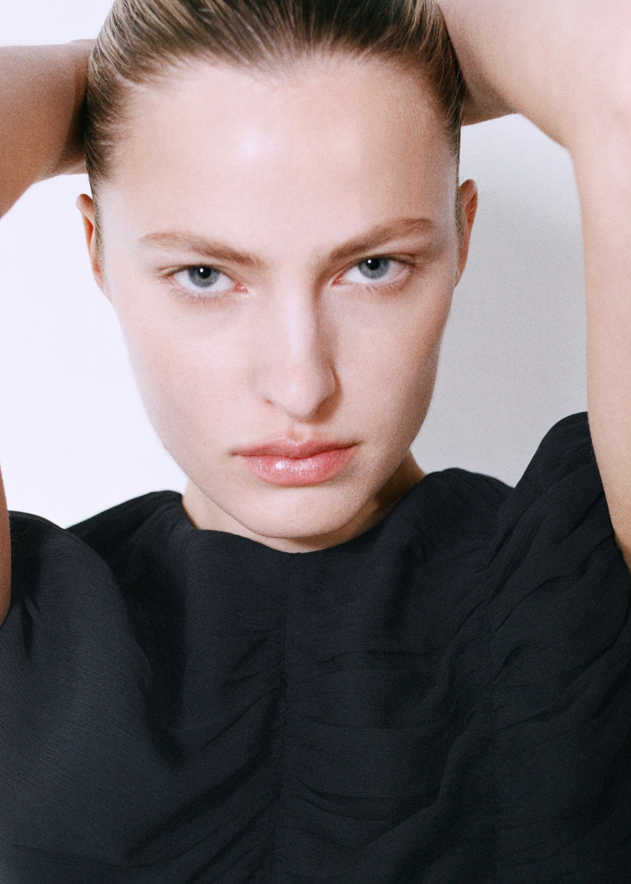 A person with slicked-back hair and blue eyes, wearing a black top, looks directly at the camera with a serious expression, arms raised.