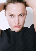 Placeholder for: A person with slicked-back hair and blue eyes, wearing a black top, looks directly at the camera with a serious expression, arms raised.