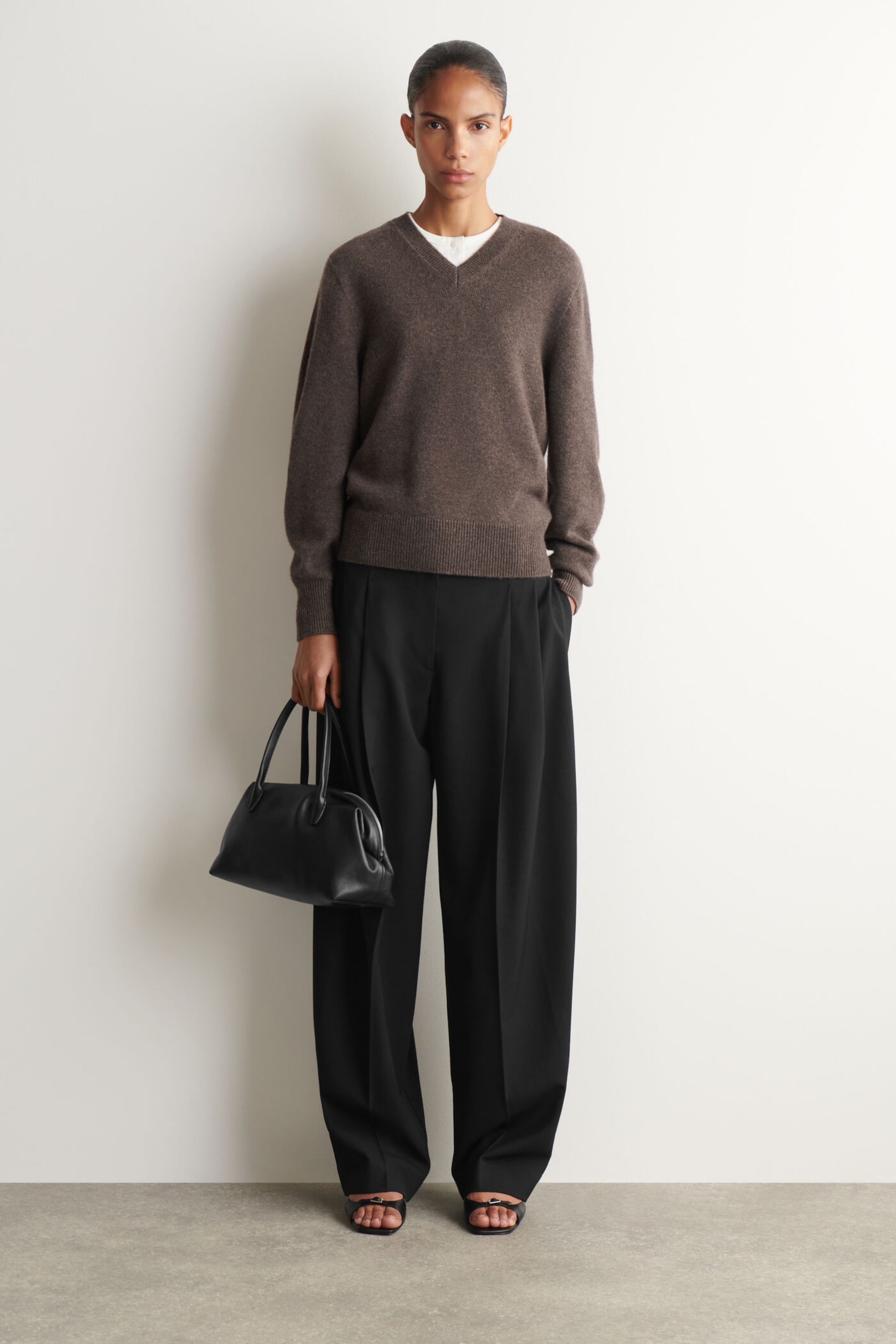 Person in a brown sweater and black pants, holding a black handbag, standing against a plain white background.