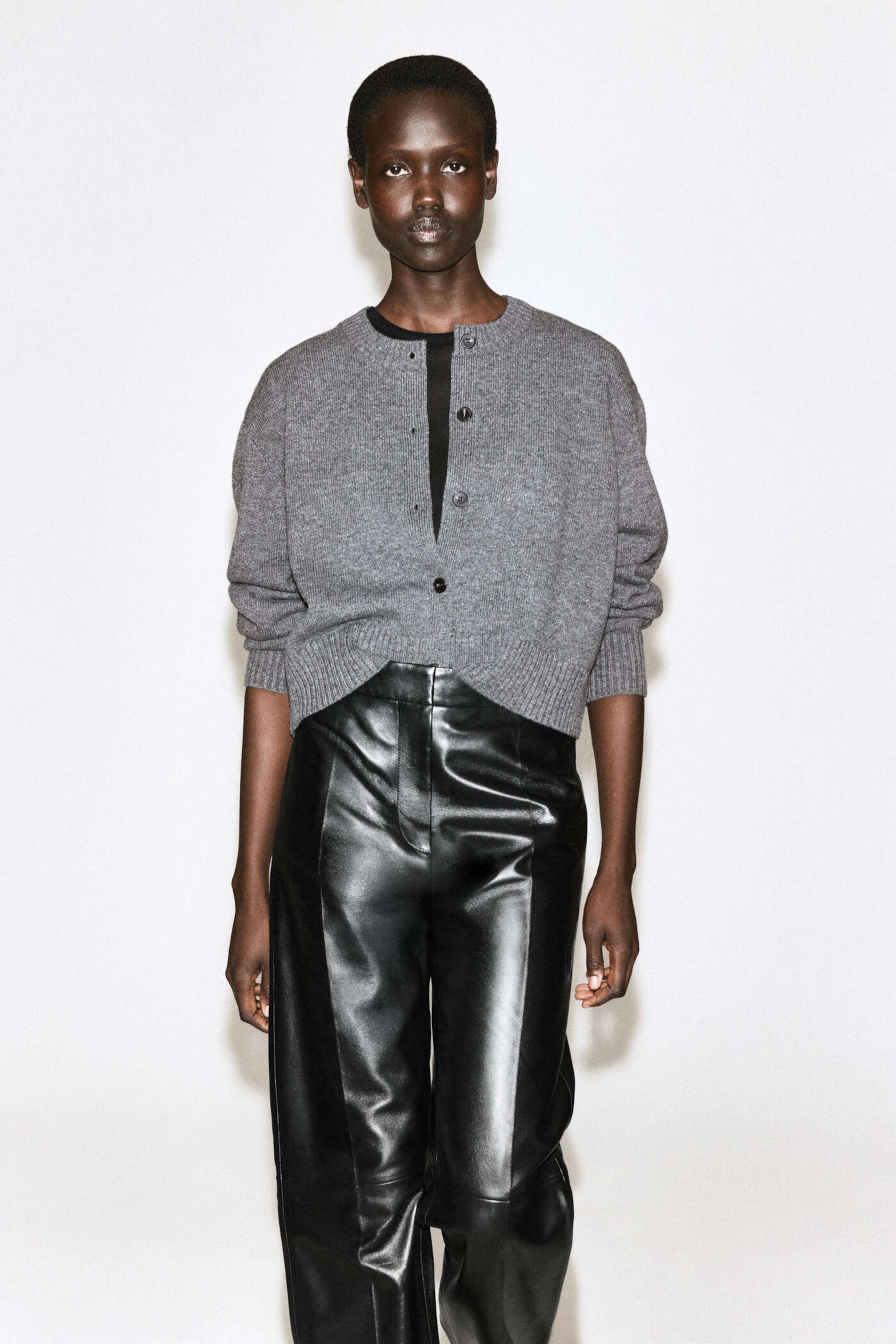 Person wearing a gray button-up sweater and black leather pants, standing against a plain white background.