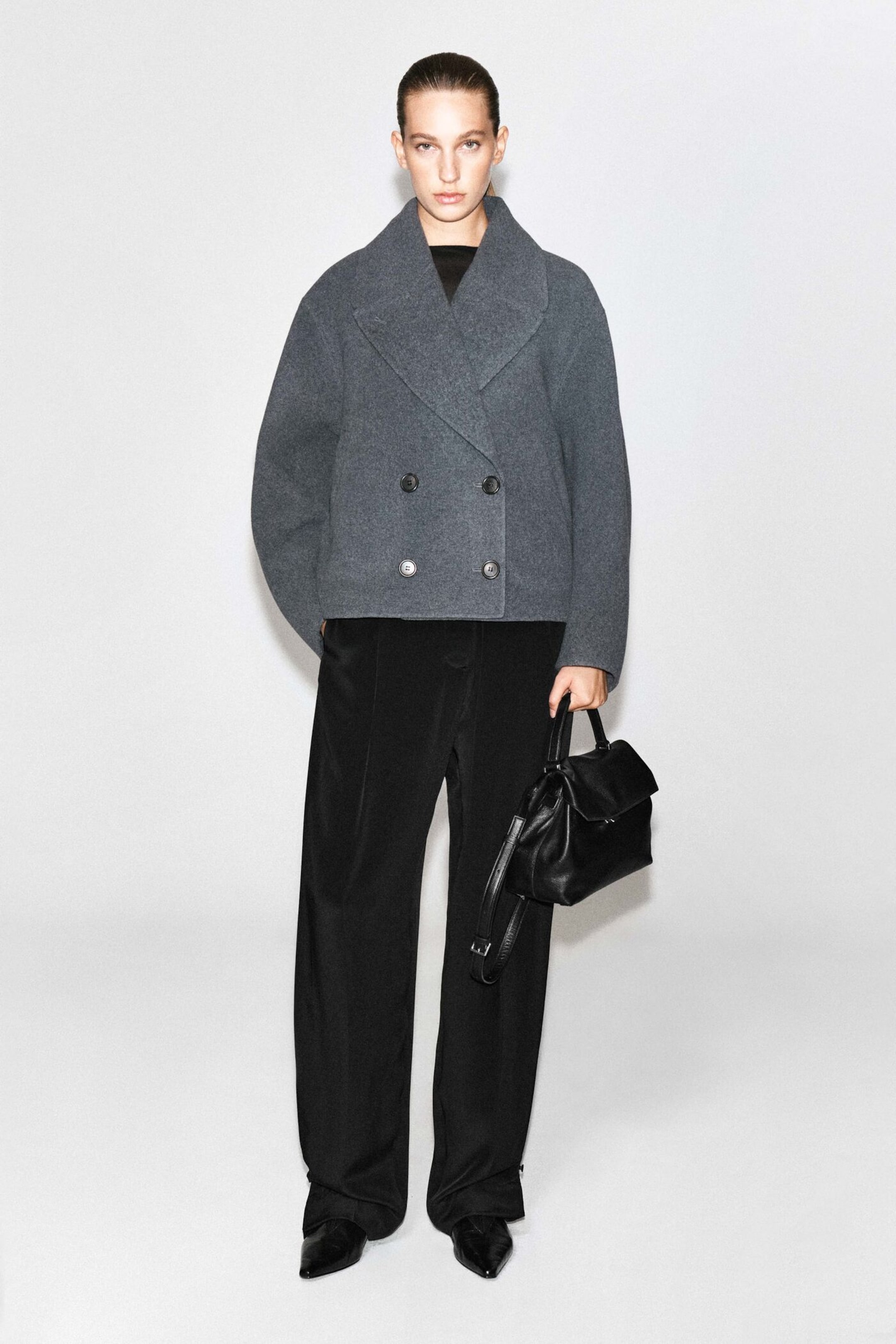 A person wearing a gray double-breasted coat and black trousers, holding a black handbag, poses against a plain background.