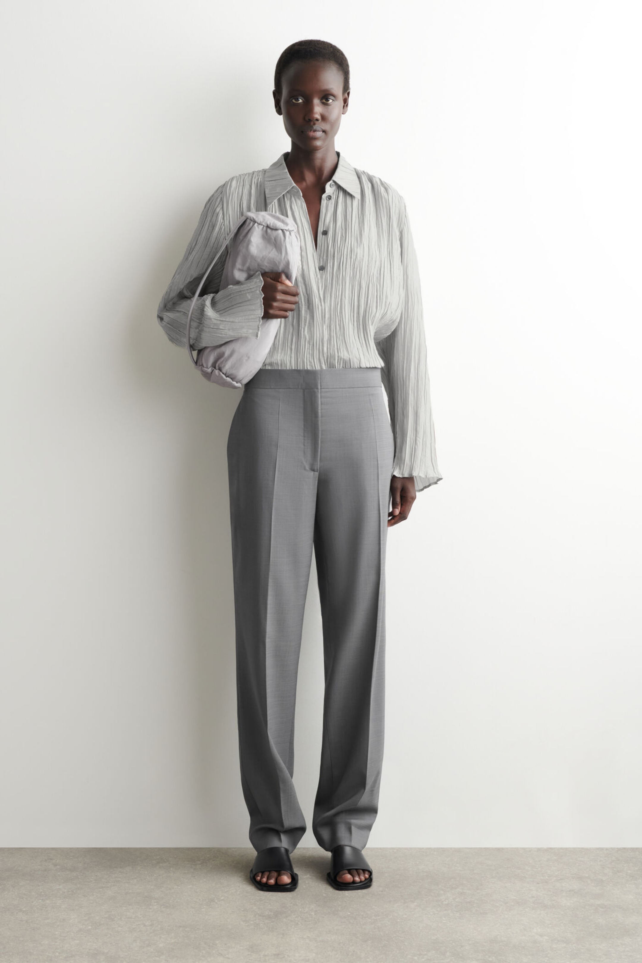 Person wearing a light gray, pleated shirt with gray trousers, holding a matching bag, standing against a plain white background.