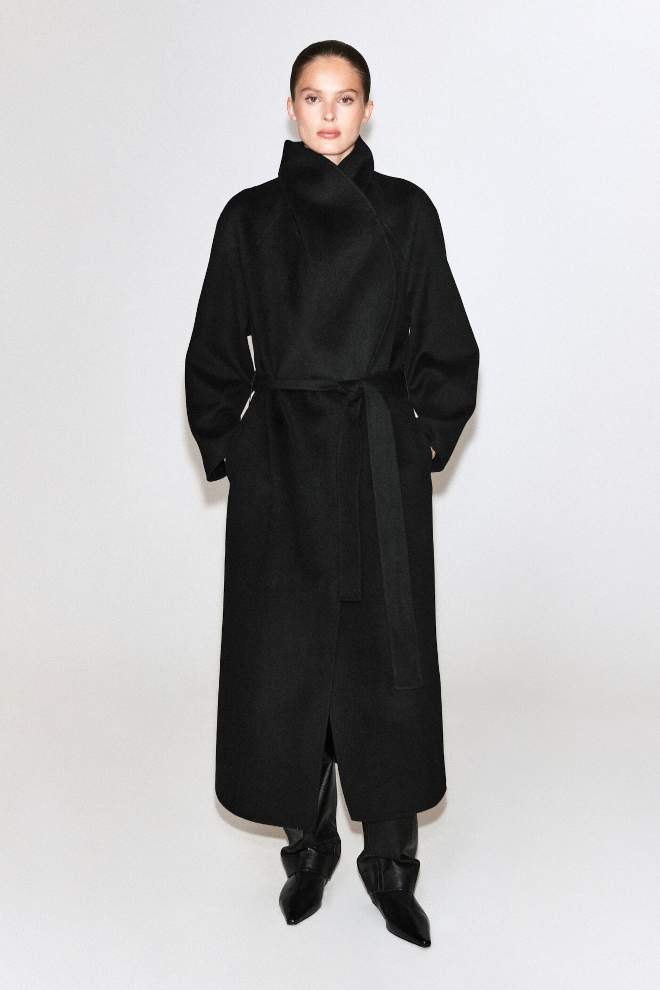 A person wearing a long, black wrap coat with a belt, standing against a plain white background.