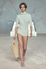 Placeholder for: Model in a cream knit outfit with a collar and shorts, carrying a large beige tote, walks on a minimalist runway with a marble backdrop.