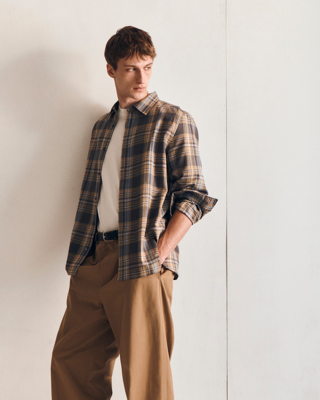Man in a plaid shirt and beige pants leans against a white wall, looking to the side with hands in pockets.