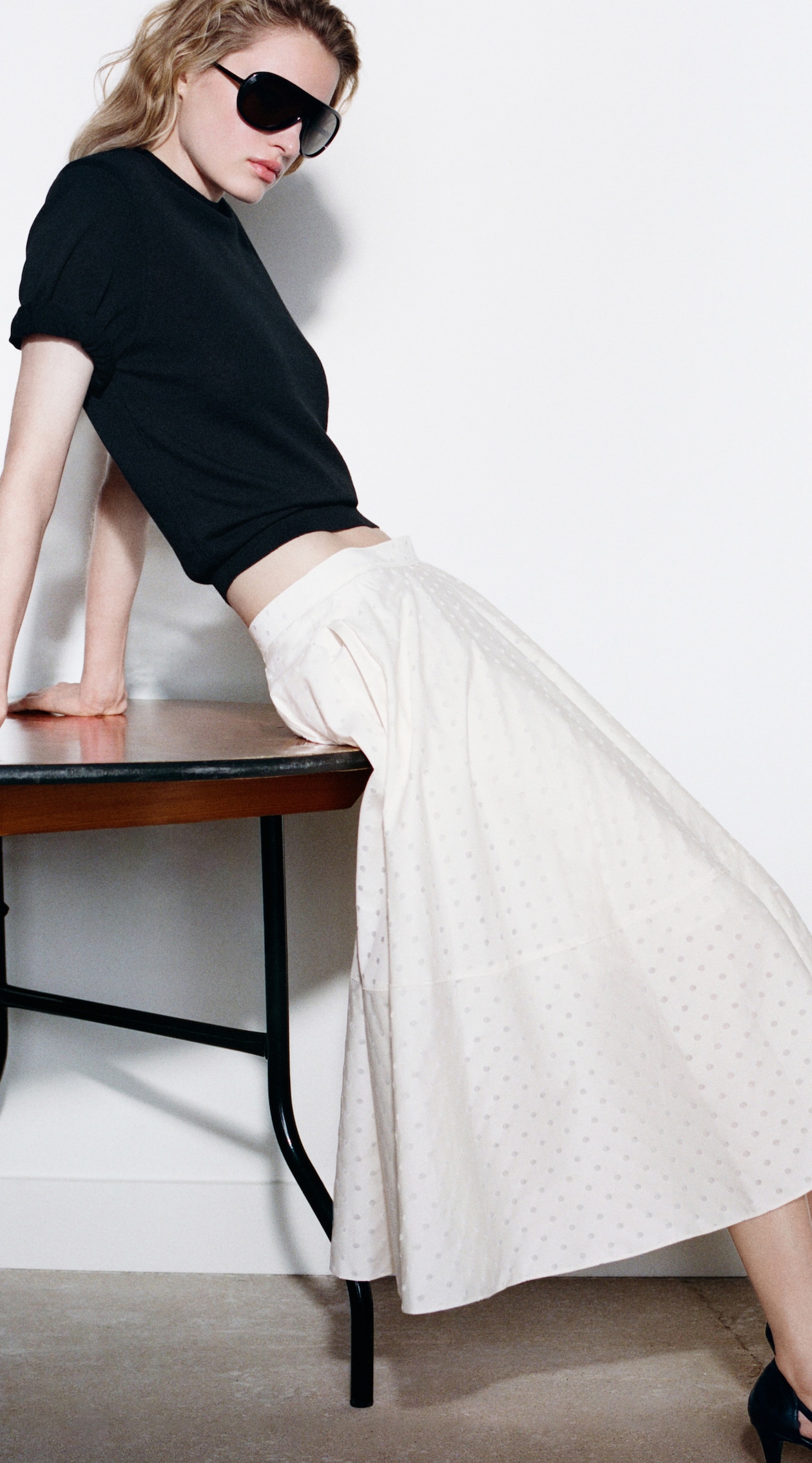 A person wearing sunglasses, a black top, and a long white skirt leans against a table in a minimalist setting.