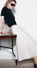 Placeholder for: A person wearing sunglasses, a black top, and a long white skirt leans against a table in a minimalist setting.
