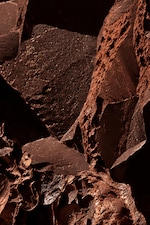 Placeholder for: Close-up of dark, textured chocolate pieces with jagged edges and cocoa dust, creating a rugged, abstract pattern.