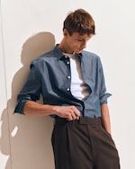 Placeholder for: Man in blue shirt with rolled-up sleeves and white undershirt adjusts brown trousers, standing against a light background.