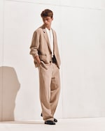 Placeholder for: Man in a beige suit and white shirt stands against a white wall, hands in pockets, looking downward pensively.