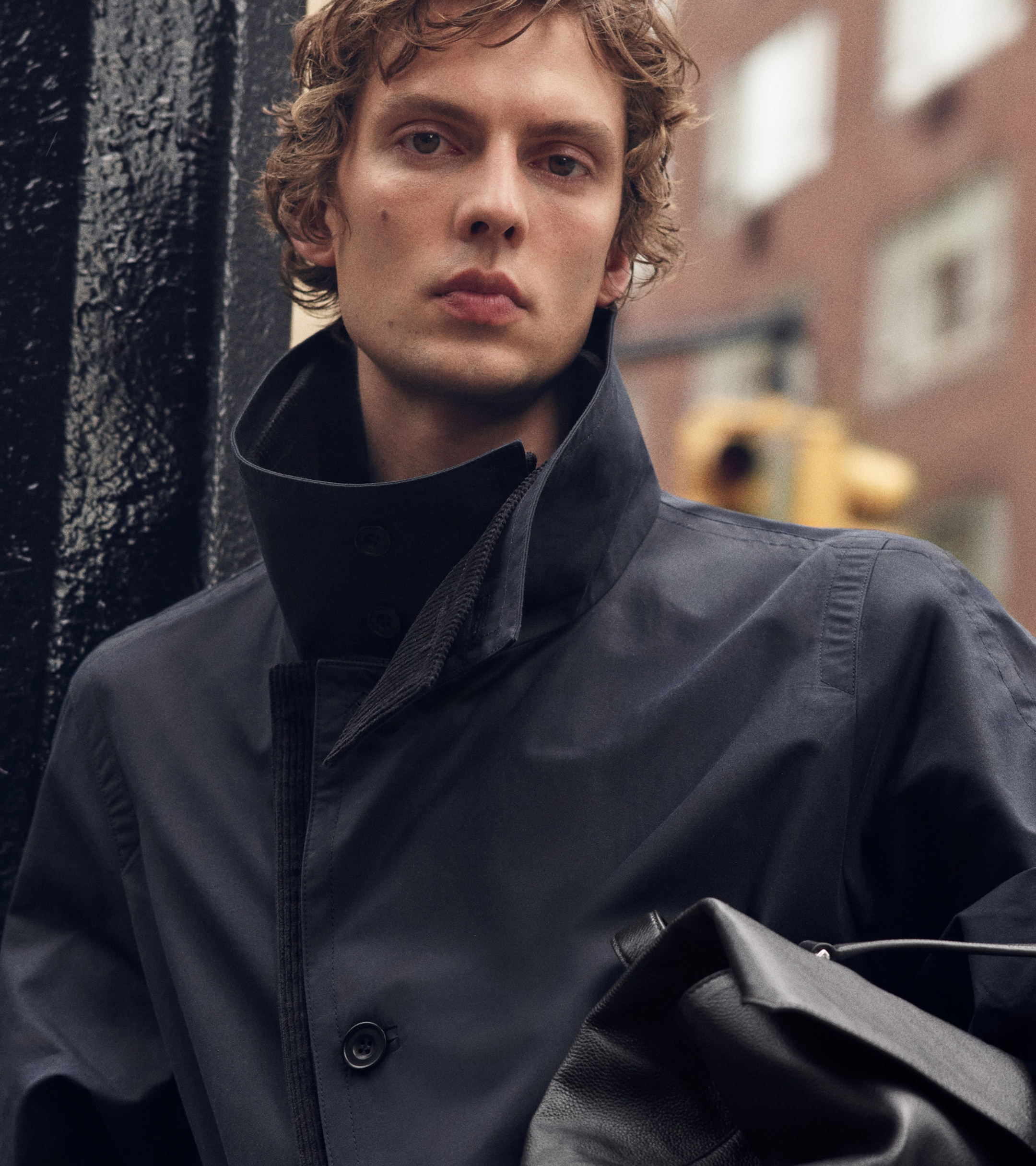 A person with curly hair wearing a high-collared black coat holds a leather bag in an urban setting.