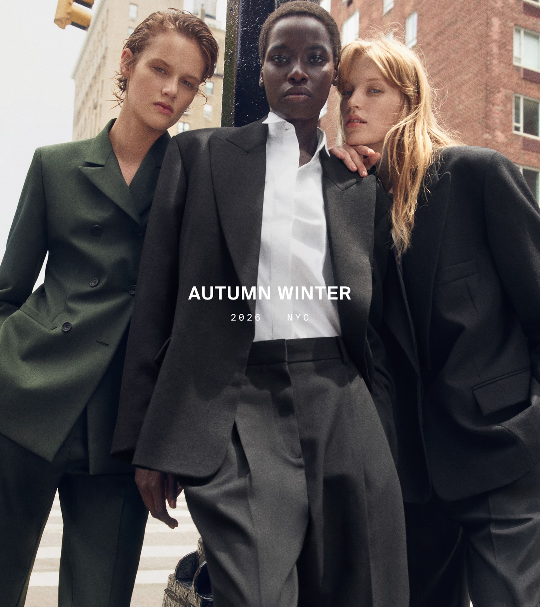 Three models in stylish suits pose confidently on a city street. Text reads "Autumn Winter 2026, NYC." Urban fashion setting.