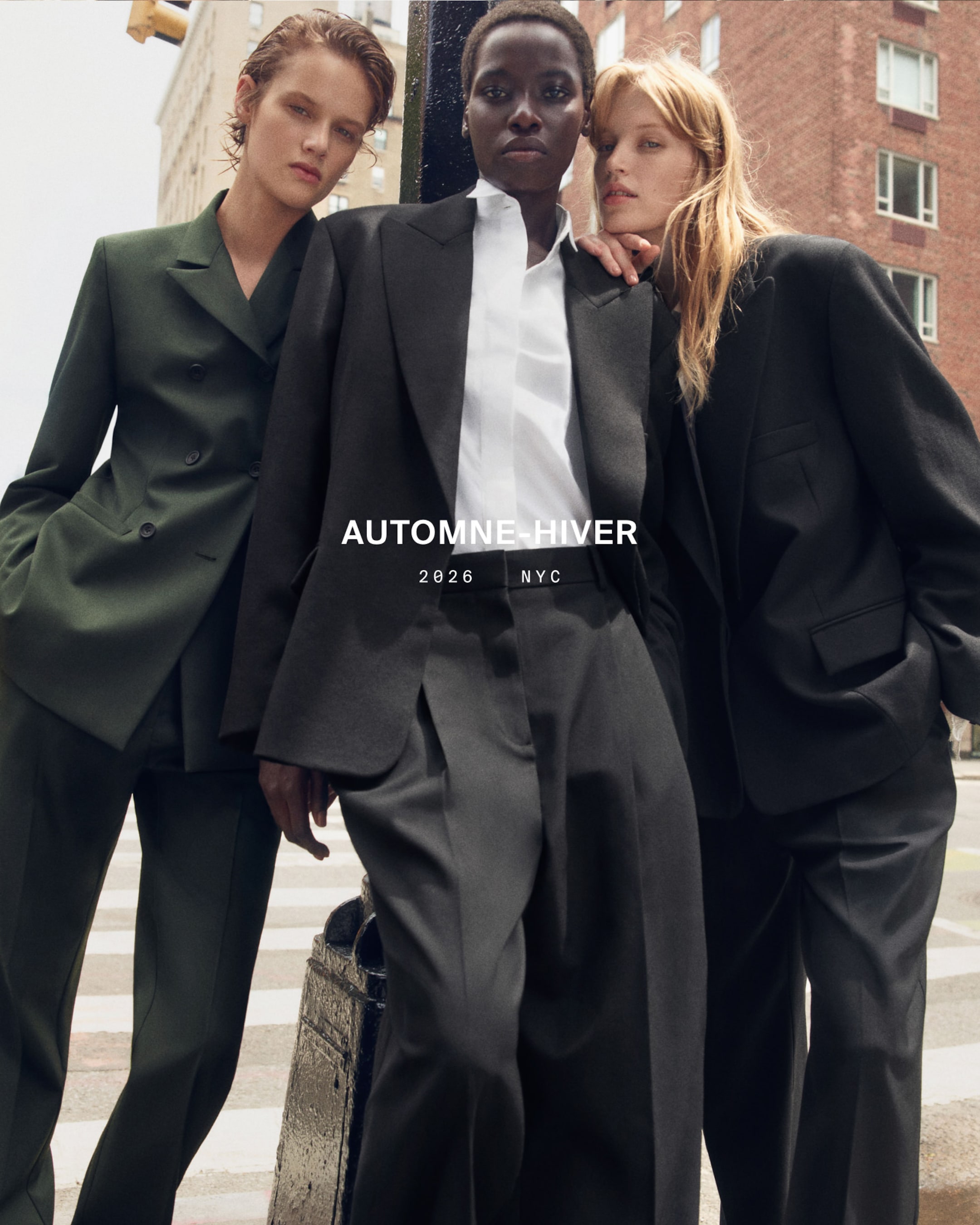 Three models in tailored suits stand on a city street, posing confidently. Text reads "AUTOMNE-HIVER 2026 NYC."