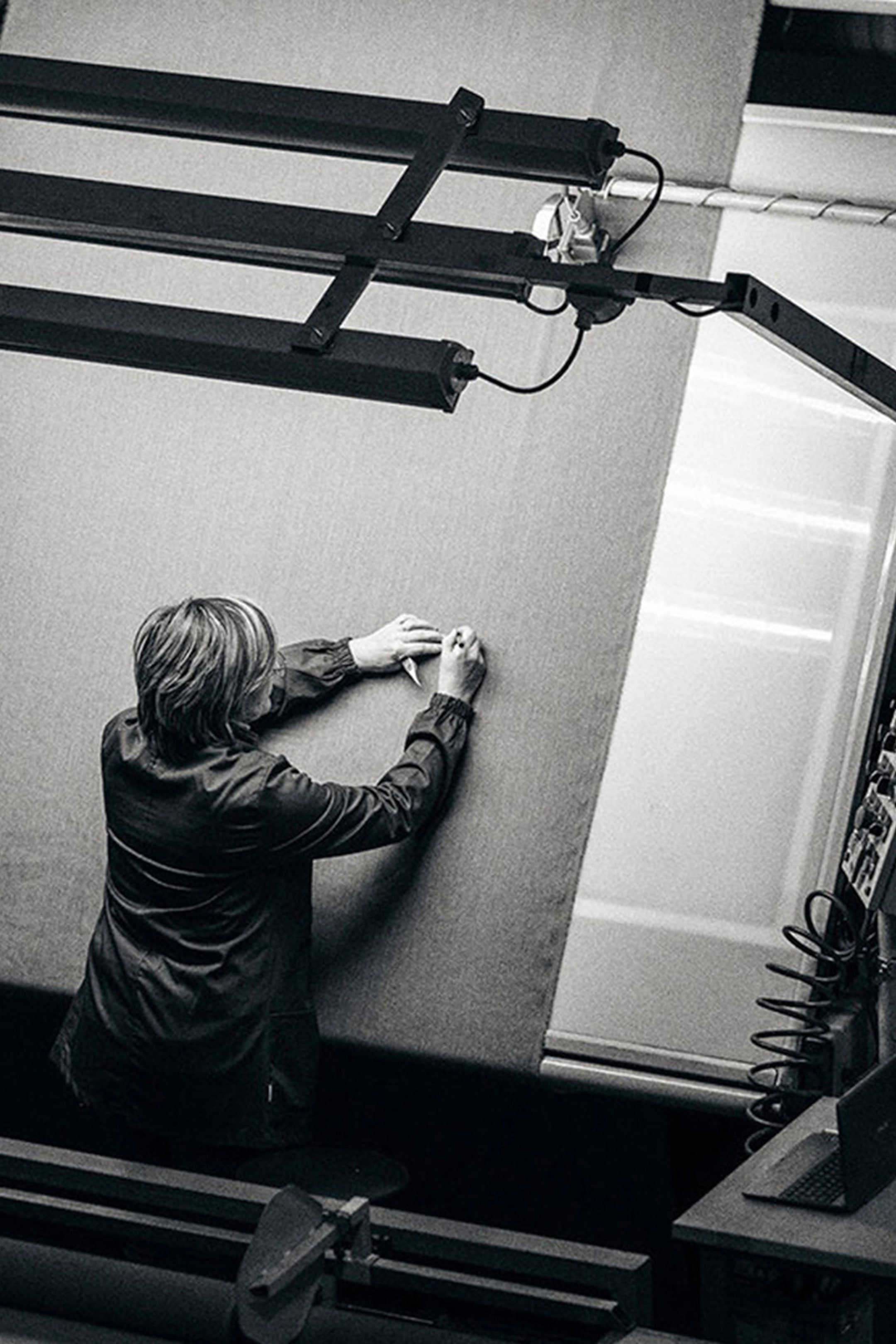 Person marking a large piece of fabric with a pen, surrounded by machinery, viewed from above in black and white.