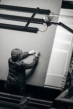 Placeholder for: Person marking a large piece of fabric with a pen, surrounded by machinery, viewed from above in black and white.