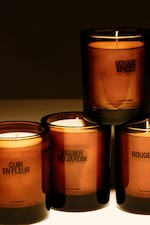 Placeholder for: Four lit candles in brown glass containers with labels: "Voyage Epices," "Cuir en Fleur," "Figuier du Jardin," and "Rouge."