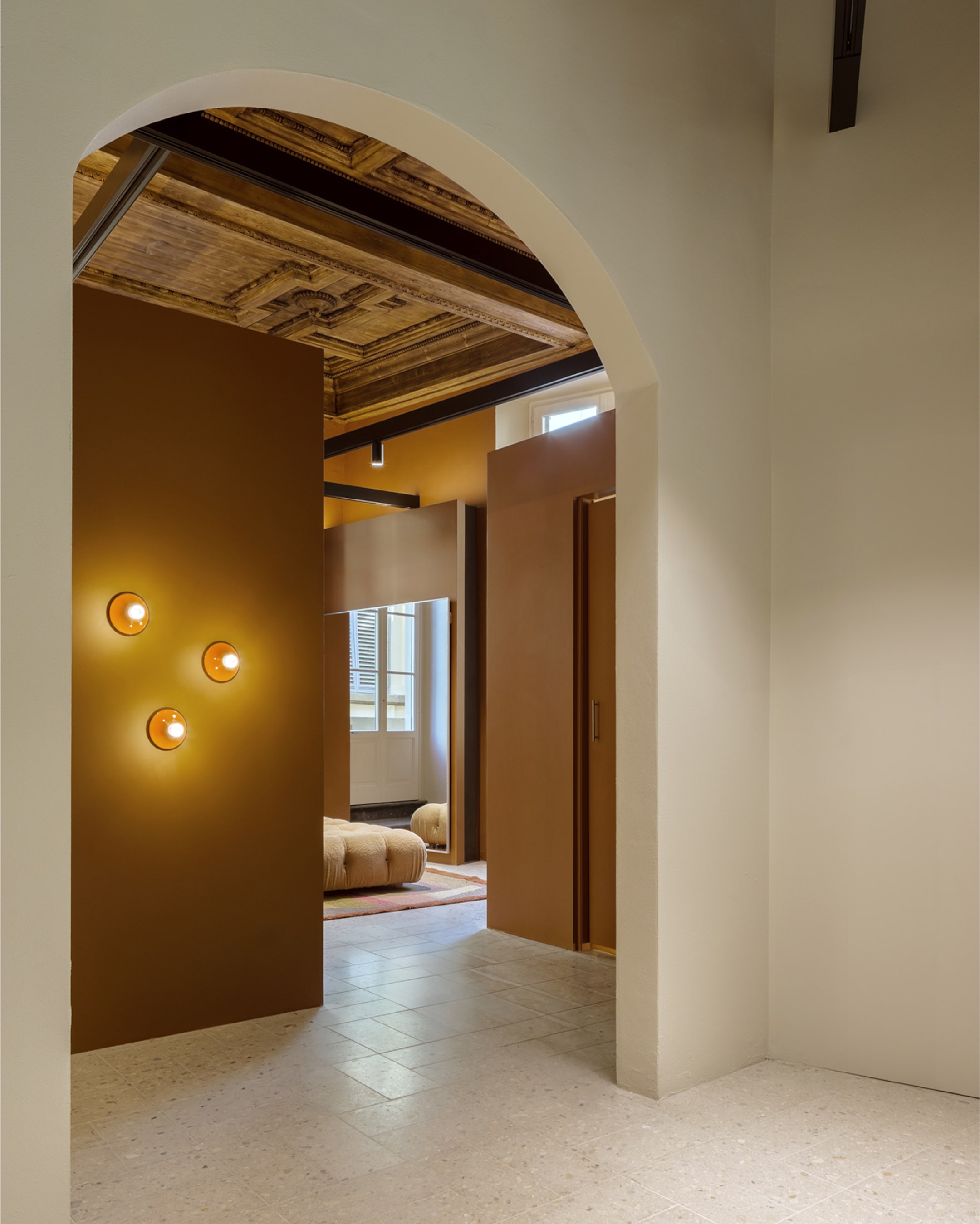 Elegant interior with arched doorway, warm brown walls, circular wall lights, and a view of a cozy seating area with natural light.