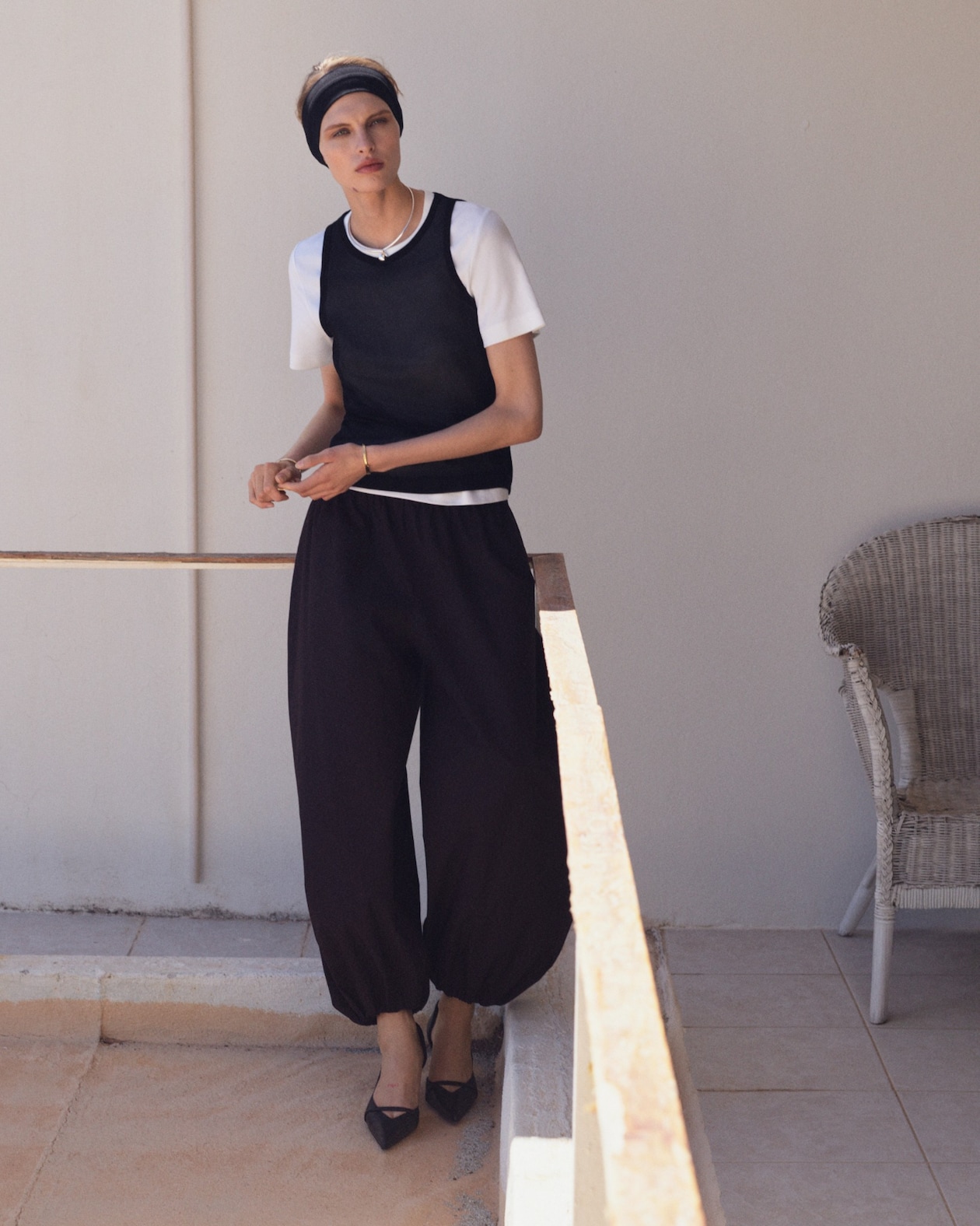 Person in a white shirt, black vest, loose black pants, and headband stands on a sunlit patio next to a white wicker chair.