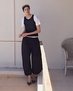 Placeholder for: Person in a white shirt, black vest, loose black pants, and headband stands on a sunlit patio next to a white wicker chair.