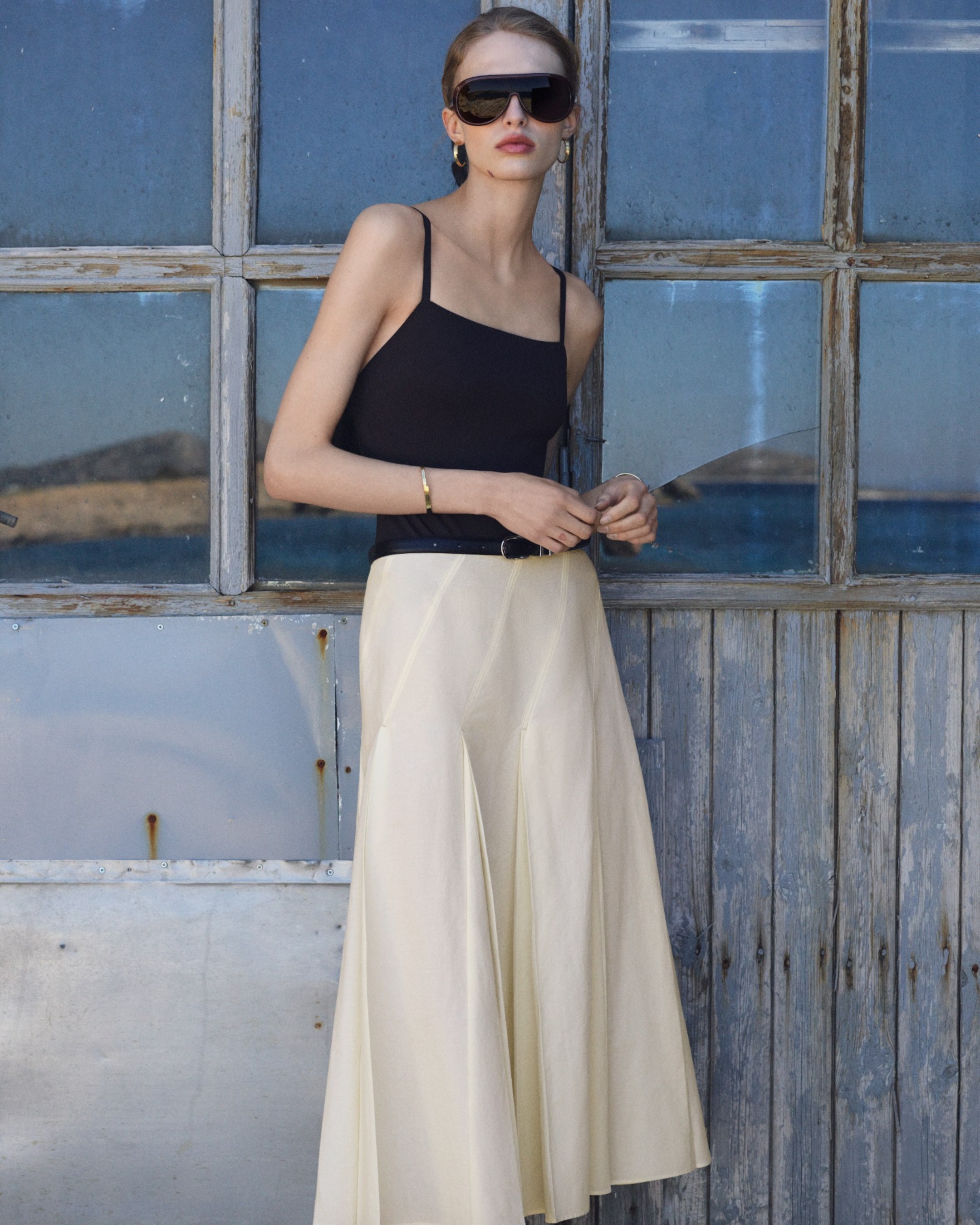 Woman in sunglasses wearing a black tank top and cream skirt, standing in front of a rustic wooden window with a seascape view.