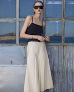 Placeholder for: Woman in sunglasses wearing a black tank top and cream skirt, standing in front of a rustic wooden window with a seascape view.