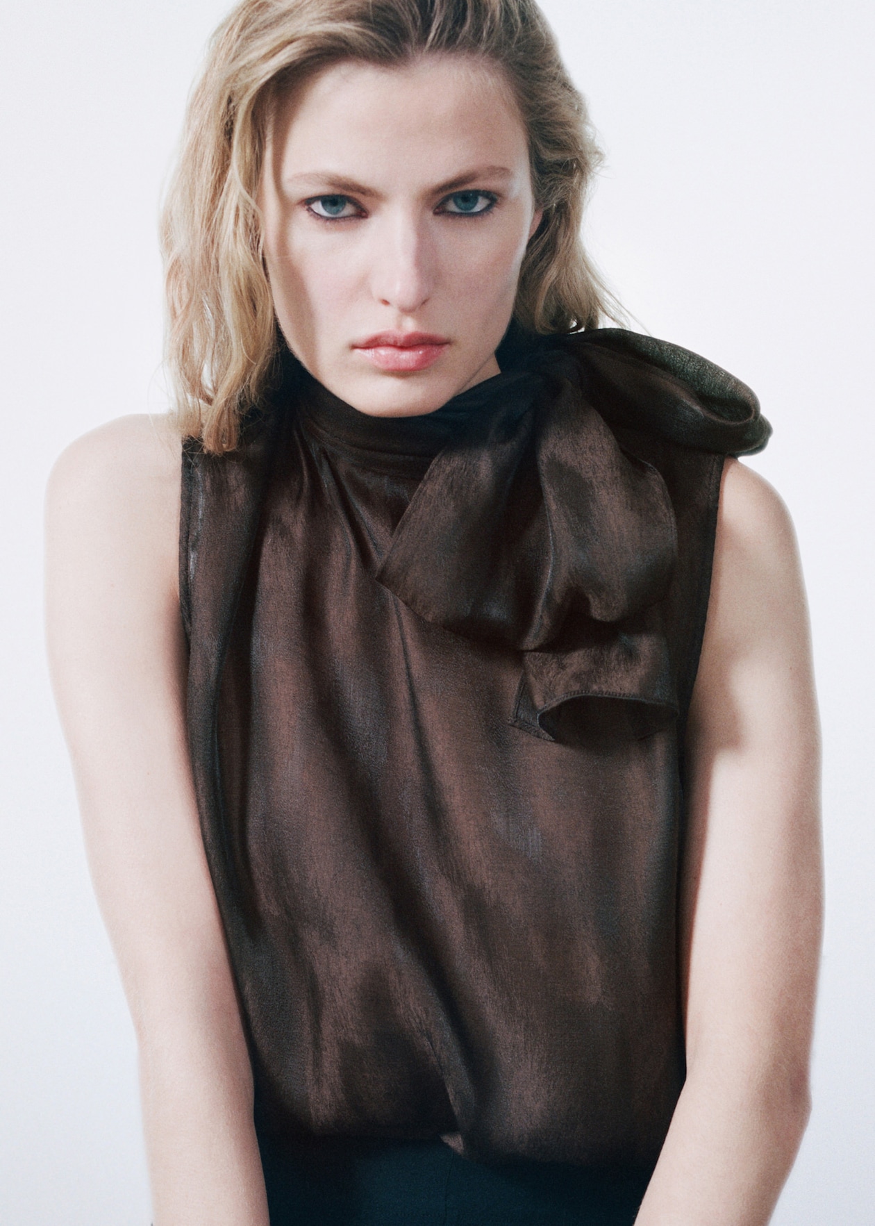 A person with blonde hair wearing a dark sheer top with a large bow, looking directly at the camera against a neutral background.