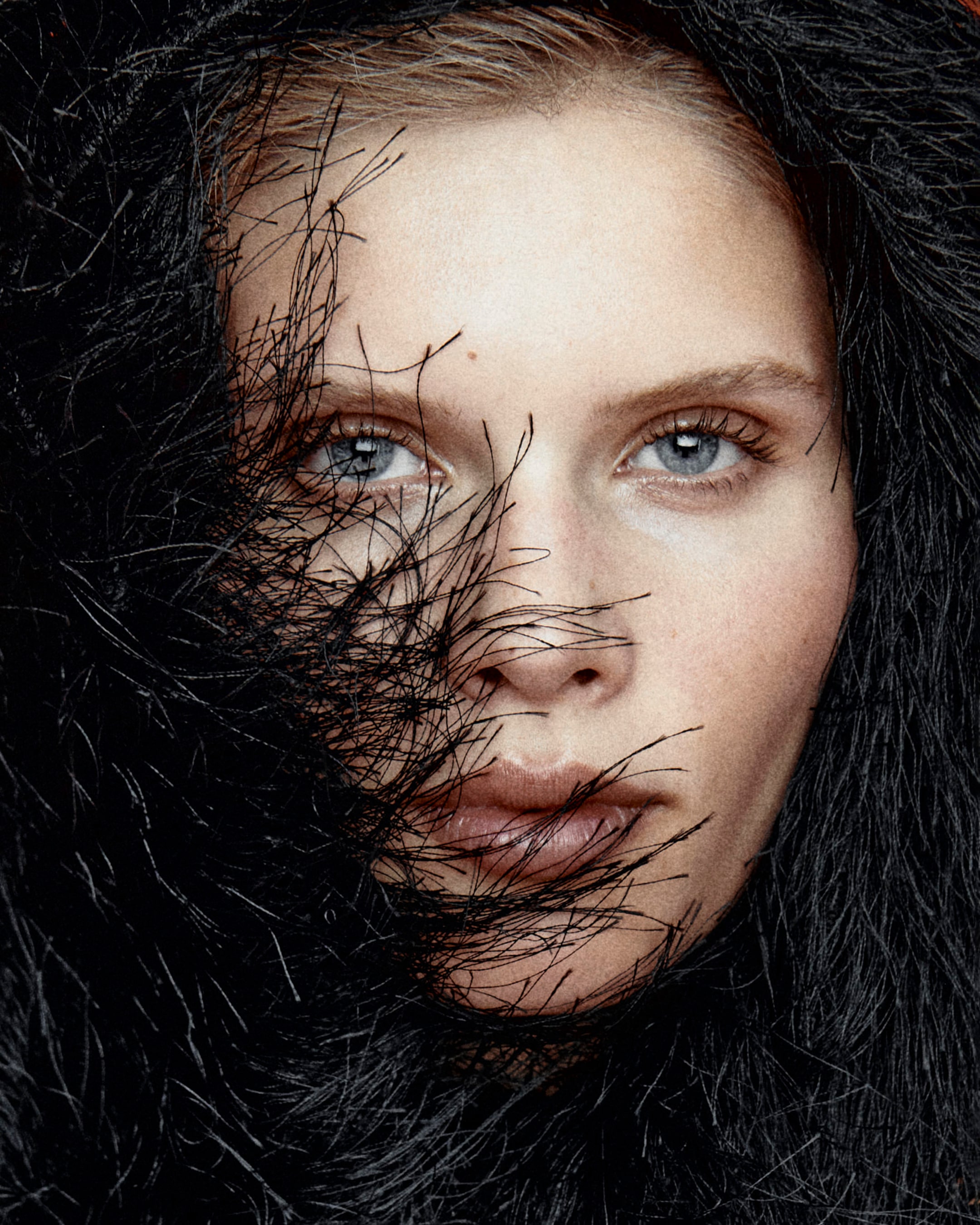 Close-up of a woman's face partly veiled by black feathery strands, with piercing blue eyes and natural lips.