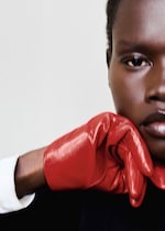 Placeholder for: Close-up of a person's face with their chin resting on a hand wearing shiny red gloves, against a neutral background.