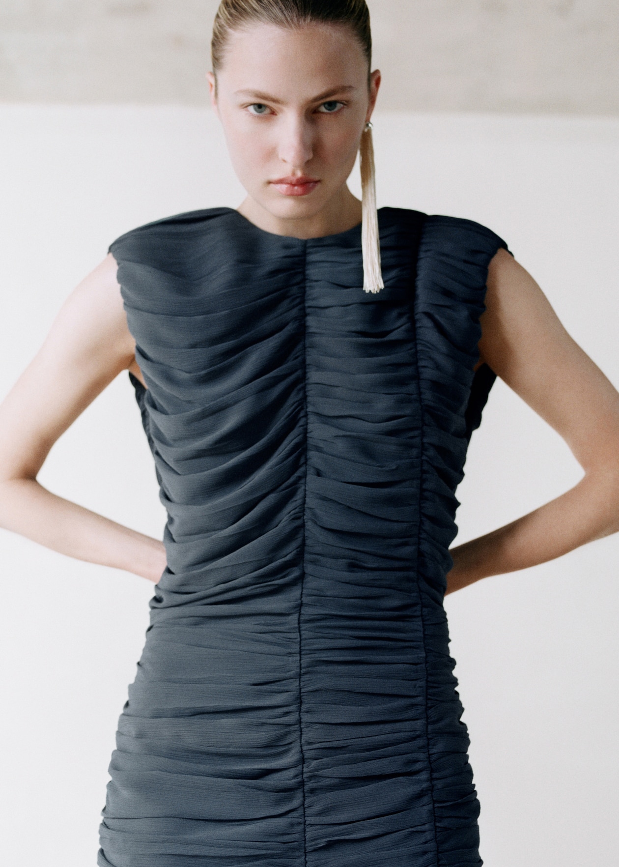 Person wearing a sleeveless, ruched black dress, with a serious expression and long tassel earring, standing with hands on hips.