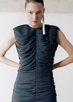 Placeholder for: Person wearing a sleeveless, ruched black dress, with a serious expression and long tassel earring, standing with hands on hips.