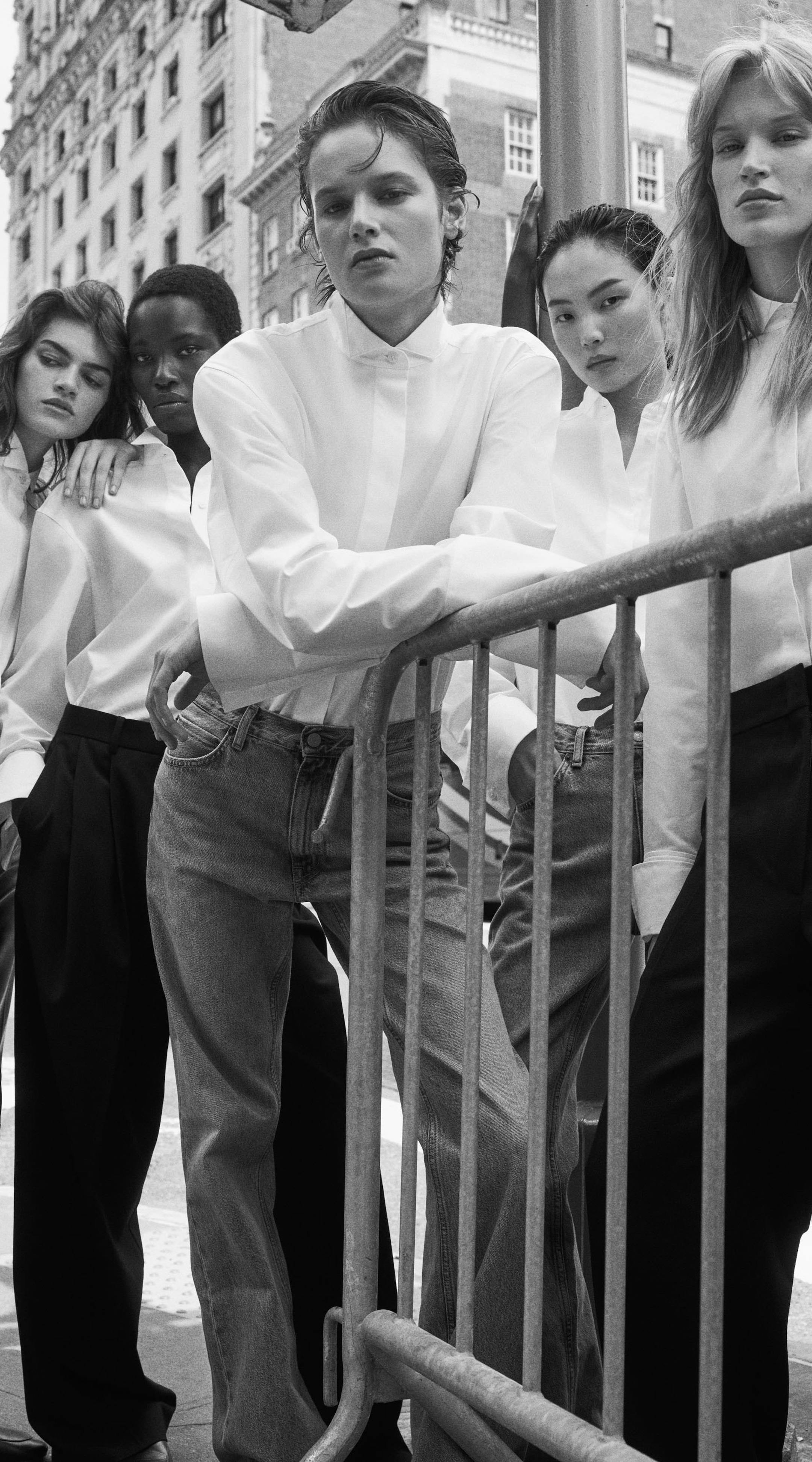 Five individuals pose in stylish white shirts and jeans, leaning against a barrier in an urban setting. Black and white photograph.