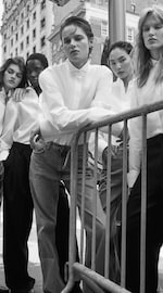 Placeholder for: Five individuals pose in stylish white shirts and jeans, leaning against a barrier in an urban setting. Black and white photograph.