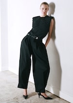 Placeholder for: Person in a stylish black outfit with wide-legged pants and pointed heels, leaning against a white wall on a textured floor.