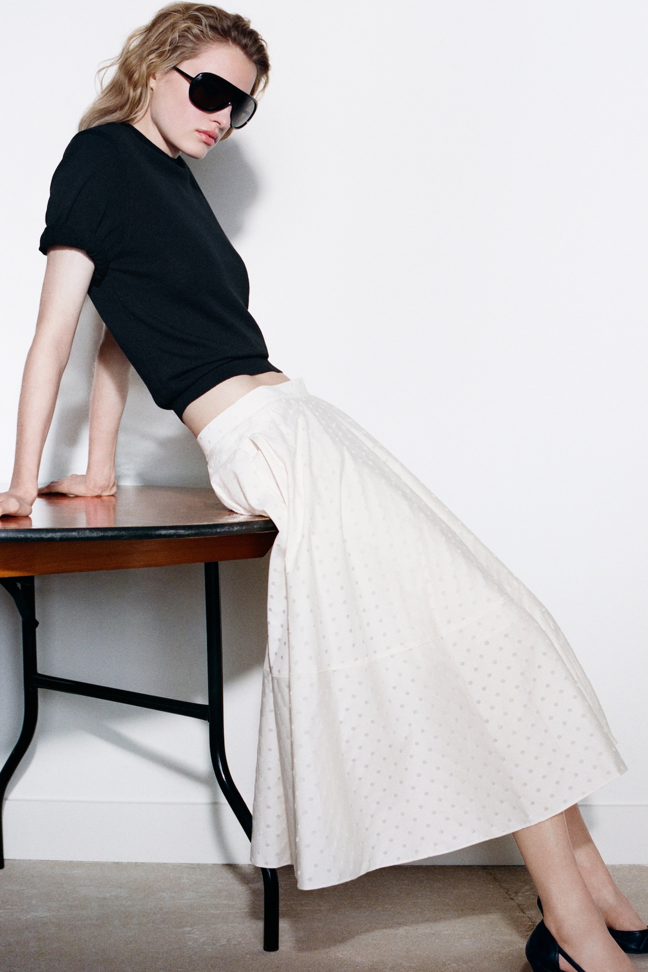Woman leaning on a table, wearing a black top, sunglasses, and a long white skirt with subtle dots, in a minimalist setting.
