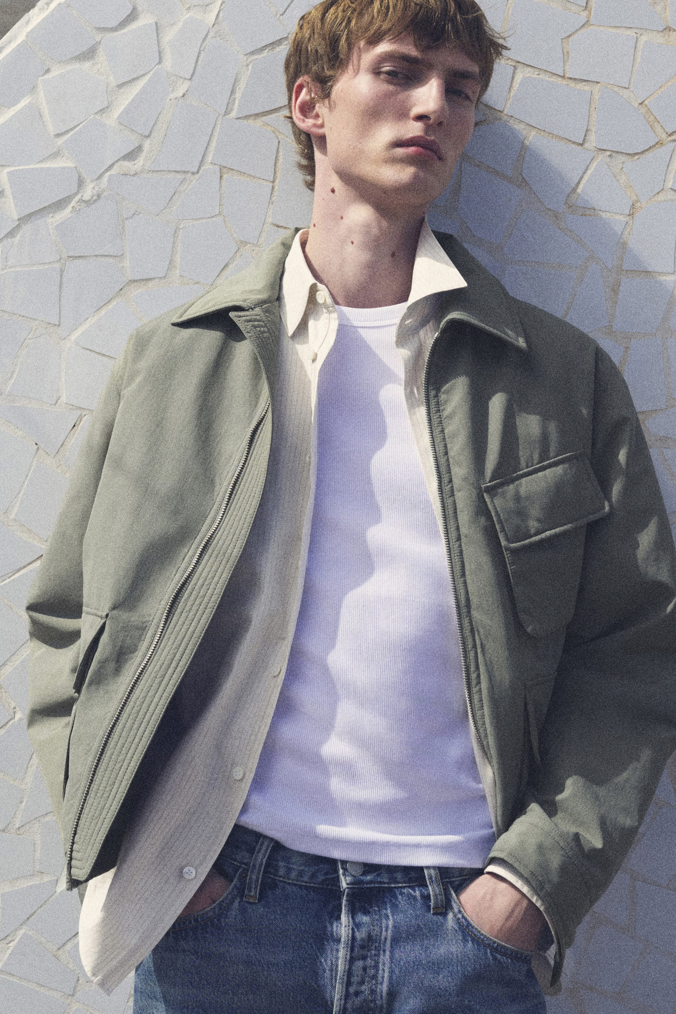 Person in a green jacket, white shirt, and jeans leans against a textured mosaic wall, looking upwards with a relaxed expression.