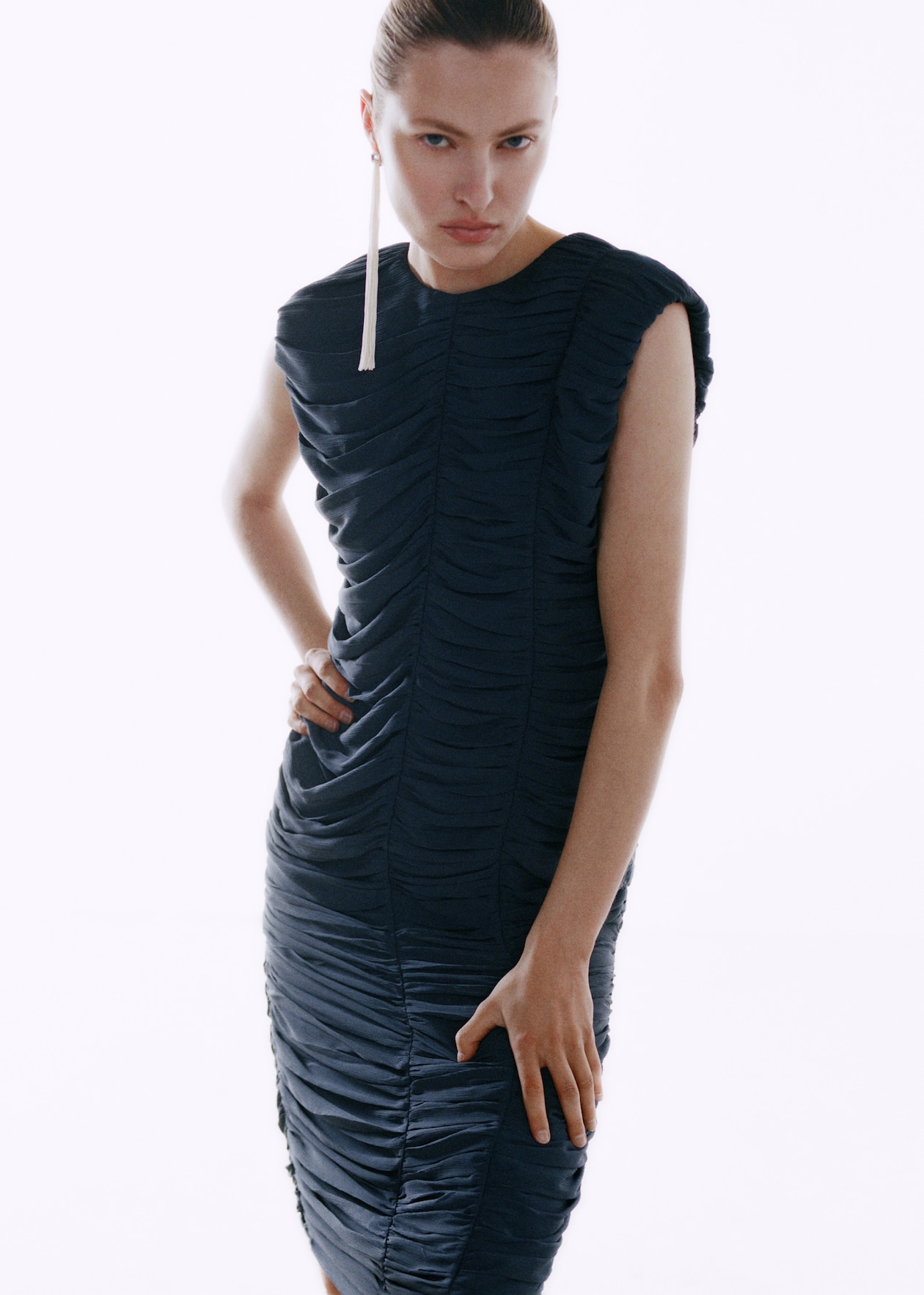 Person in a textured, ruched black dress poses with hand on hip, wearing a long, dangling earring, on a white background.