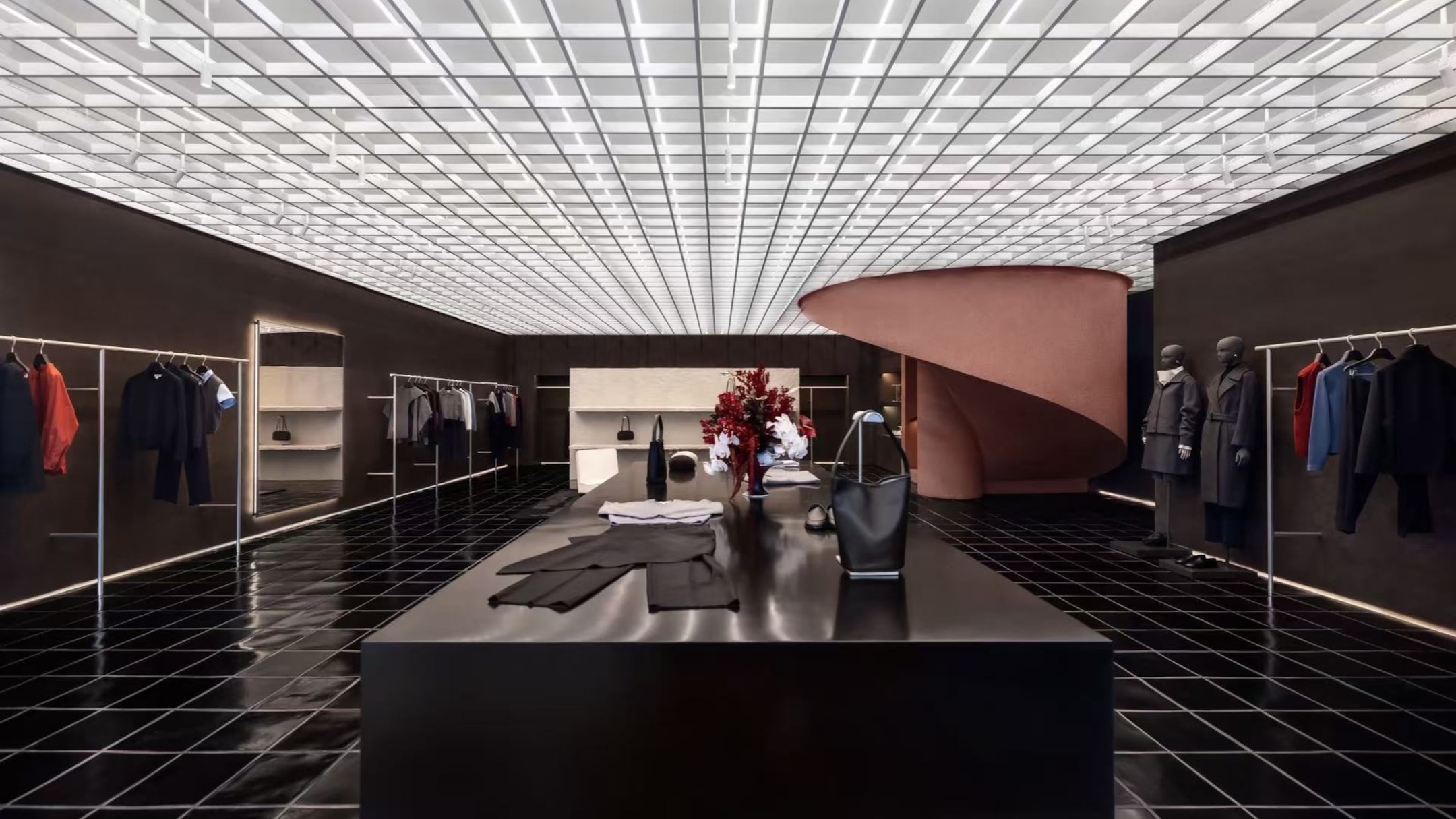 Modern clothing store interior with sleek black flooring and ceiling grid. Central display table with garments, spiral staircase, and mannequins.
