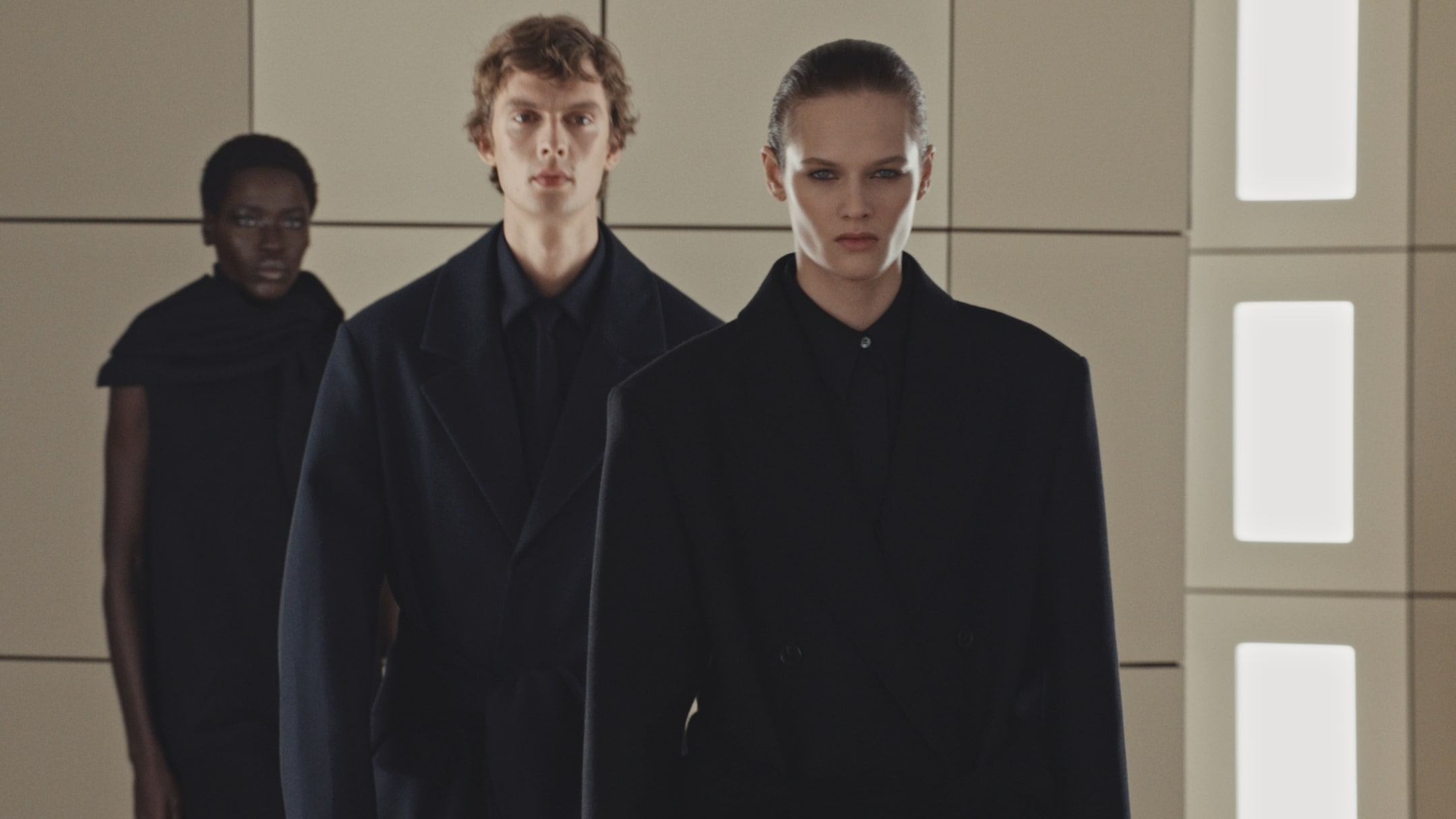 Three models in black clothing stand in a modern, brightly lit room with a geometric background.