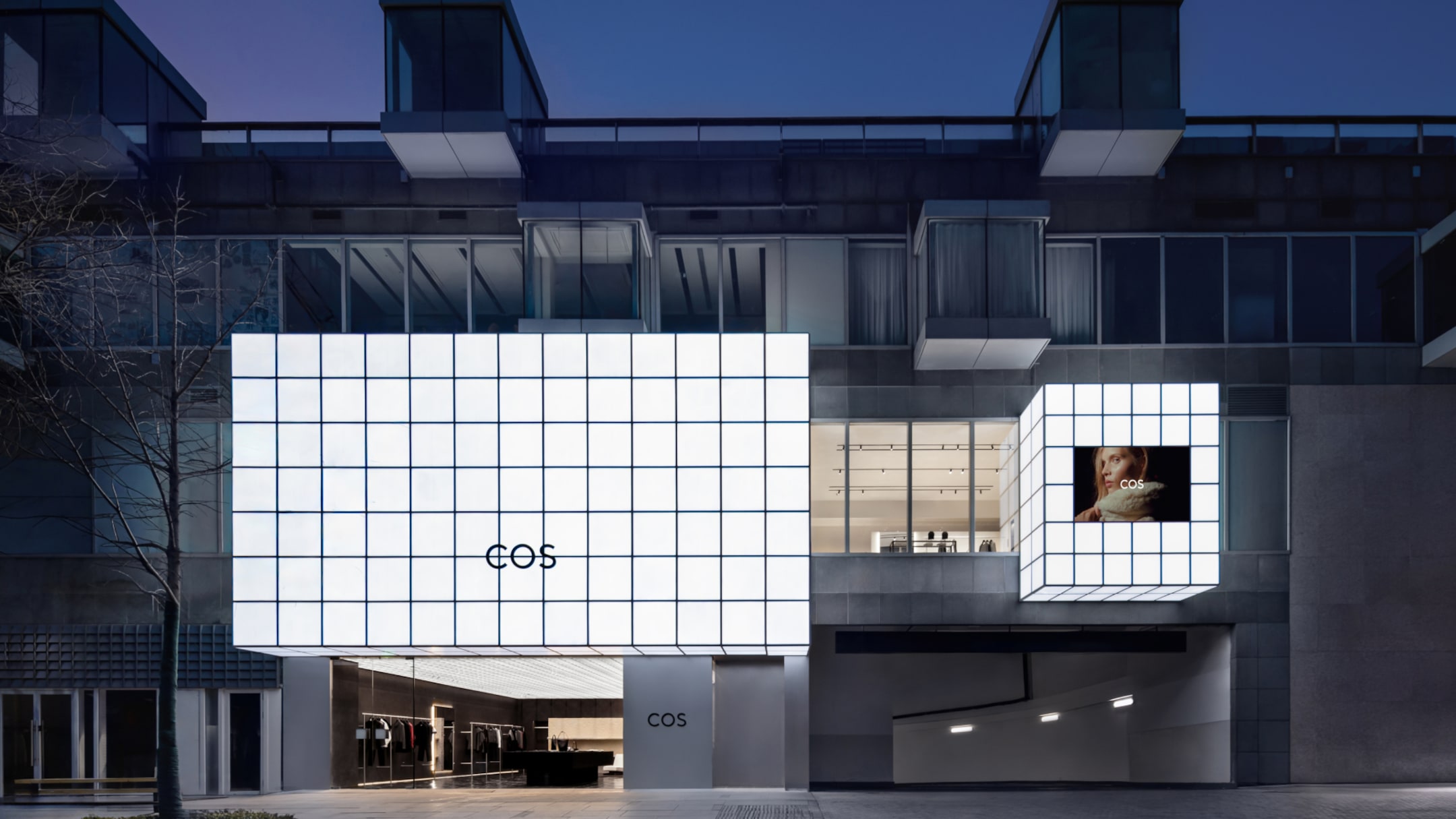 Modern building exterior with illuminated grid facade displaying "COS" logo, featuring a large digital screen and glass windows at night.