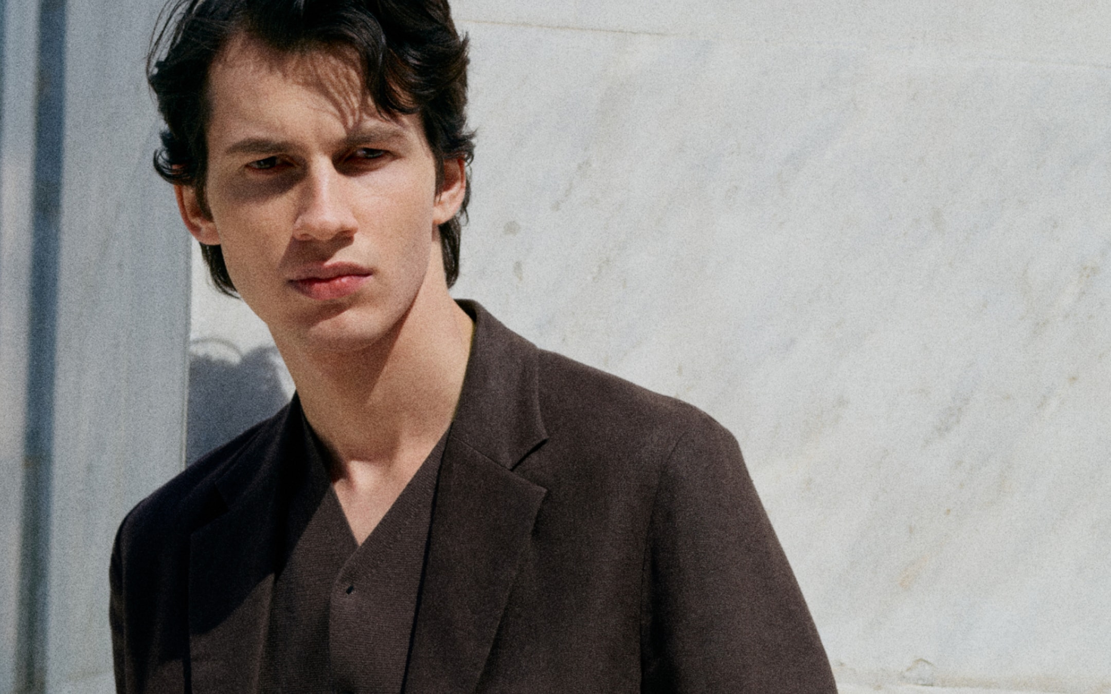 Young man in a dark blazer stands against a light stone wall, looking to the side with a thoughtful expression.