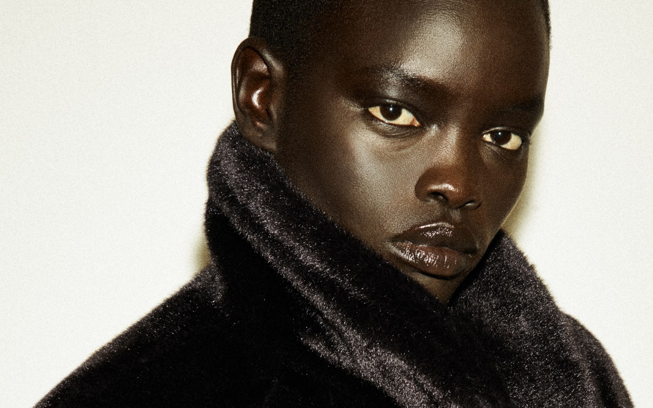 Person with short hair wearing a dark fur coat, looking directly at the camera against a plain background.