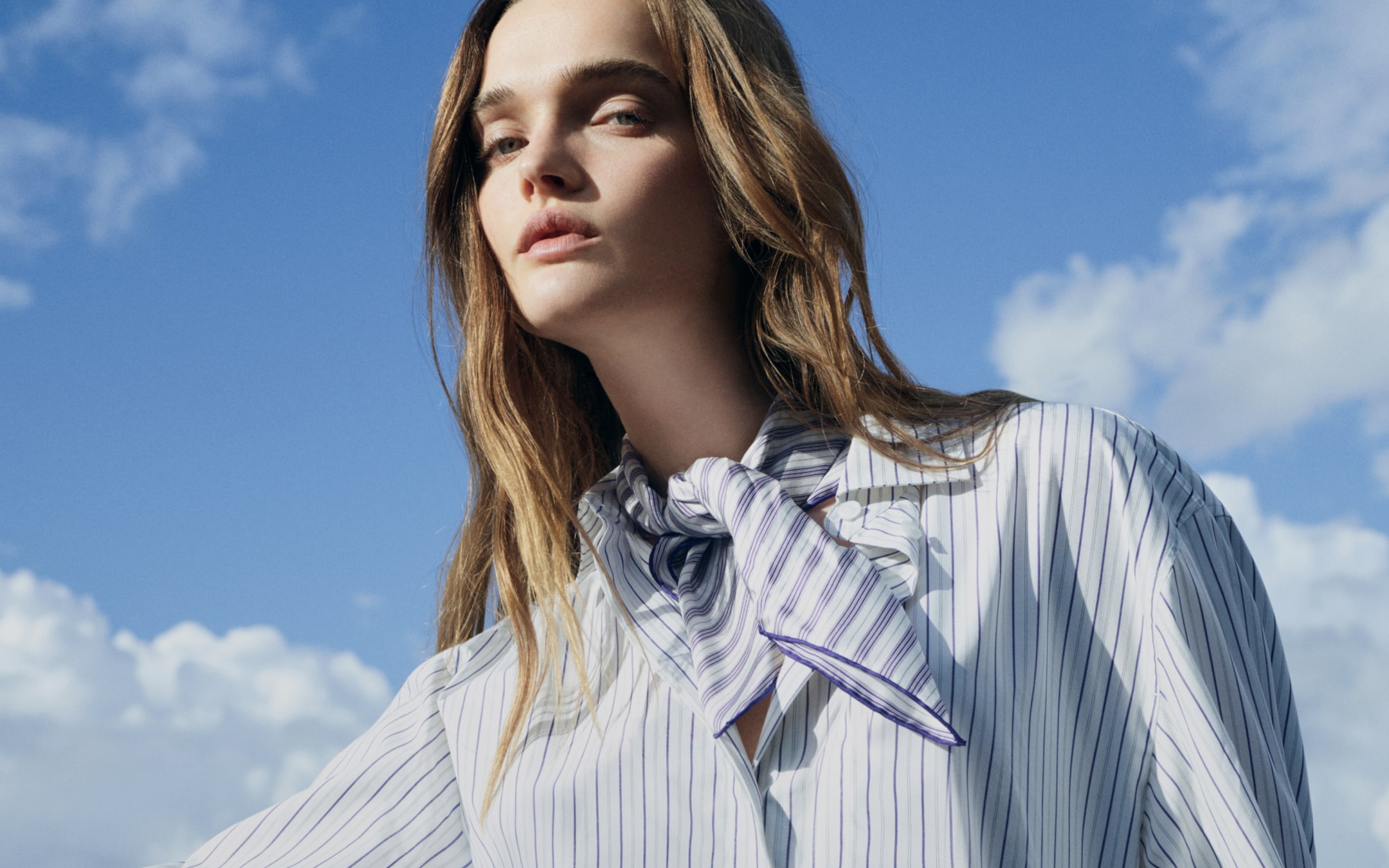Woman with long hair, wearing a striped blouse and scarf, looking upward against a blue sky with clouds.