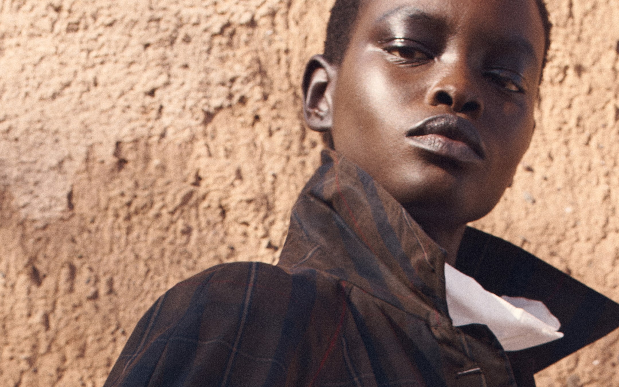 A person wearing a high-collared jacket gazes confidently to the side. The background features a textured brown wall.