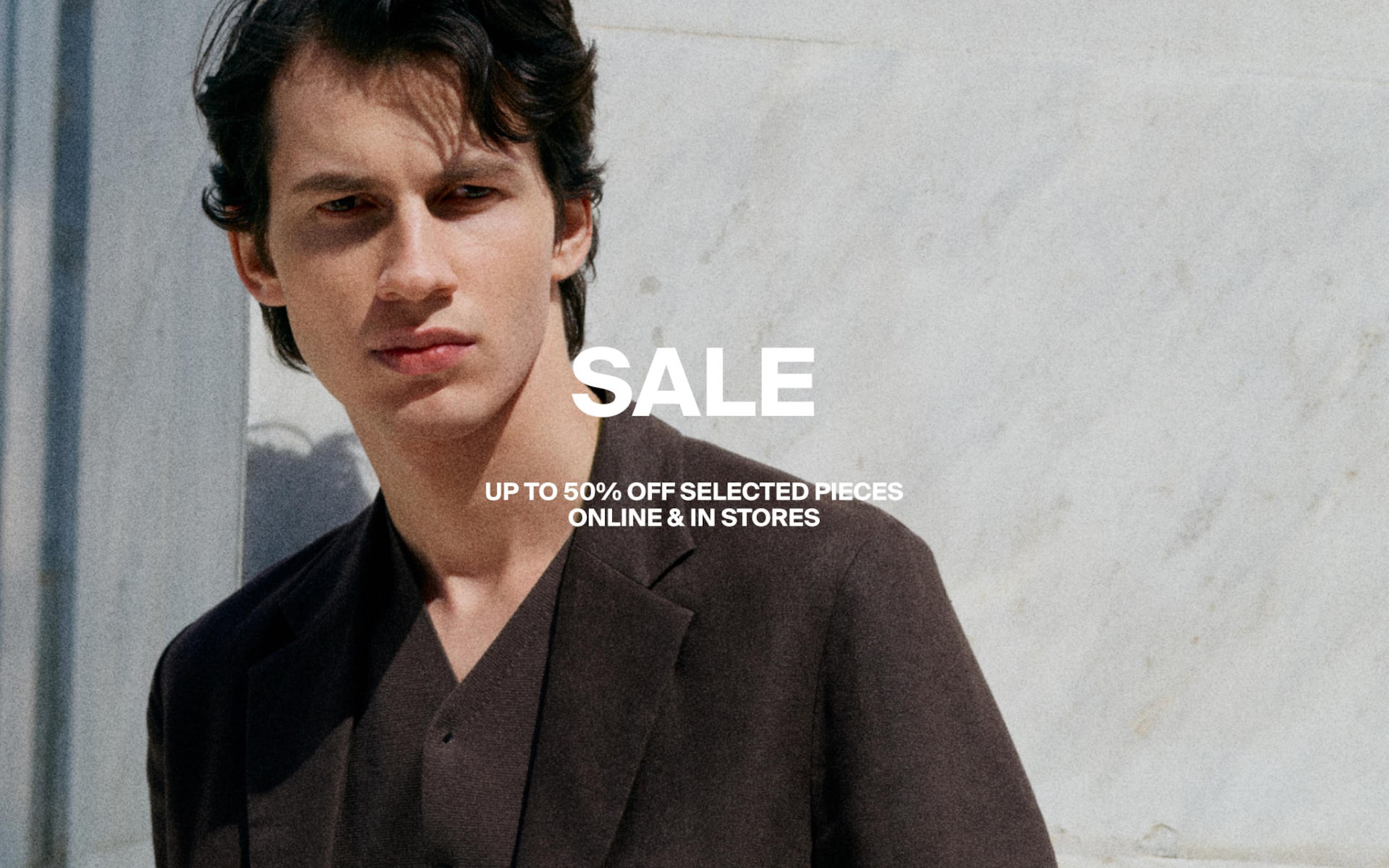 Man in a dark suit stands against a light wall; text reads "SALE: Up to 50% off selected pieces online & in stores."