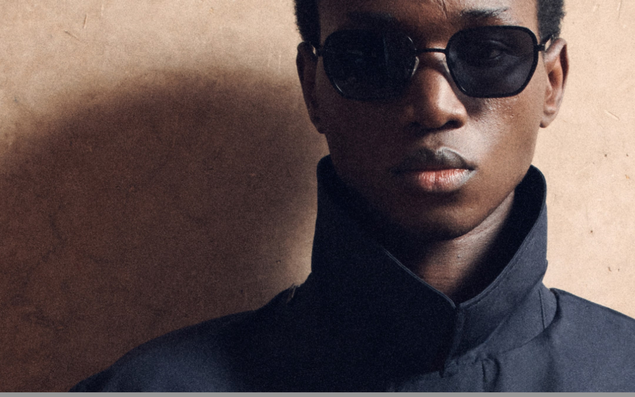 Person wearing dark sunglasses and a high-collared jacket against a textured beige background.