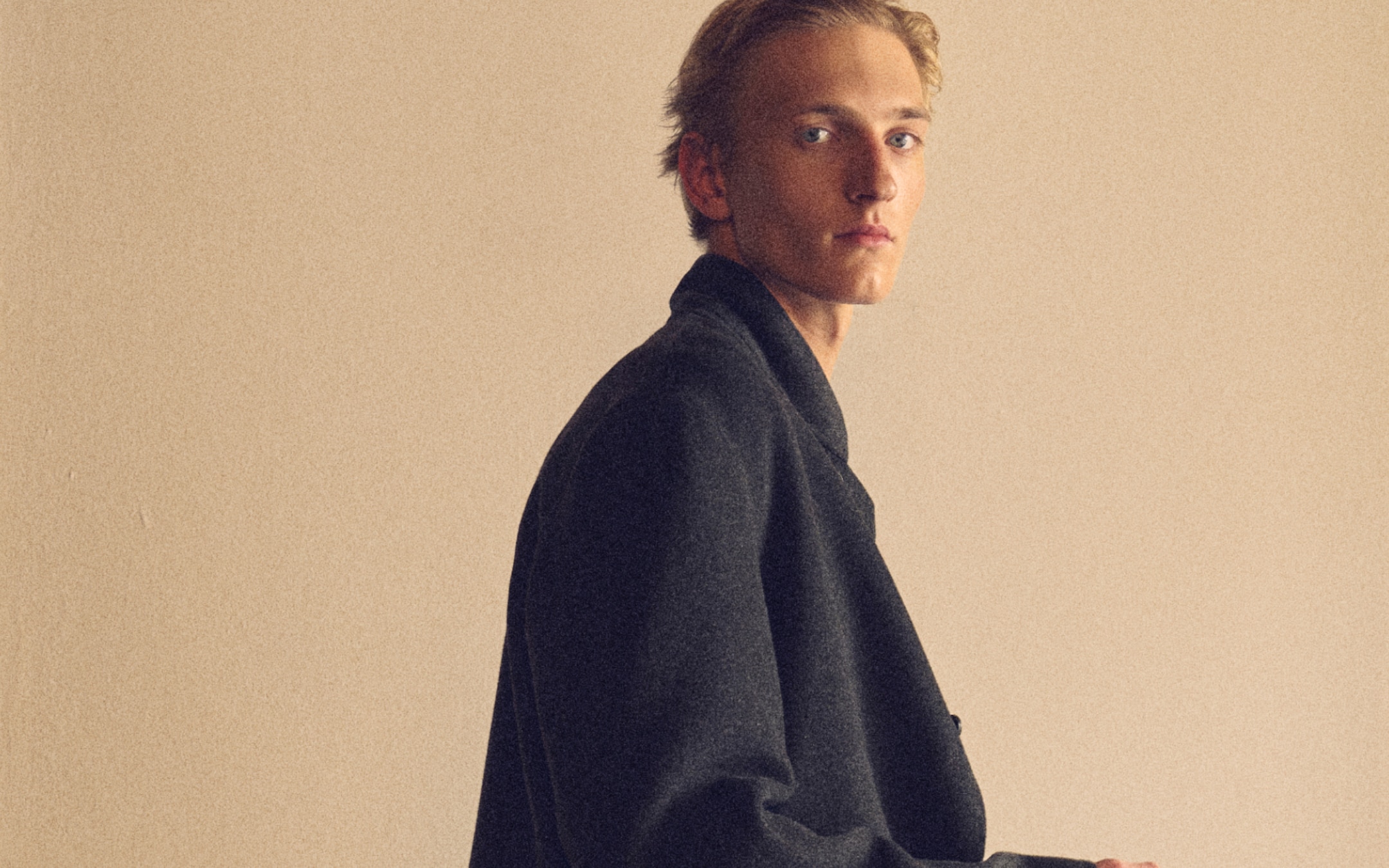 Person in a black coat looks sideways, standing against a plain beige background with a contemplative expression.