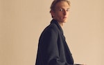 Placeholder for: Person in a black coat looks sideways, standing against a plain beige background with a contemplative expression.