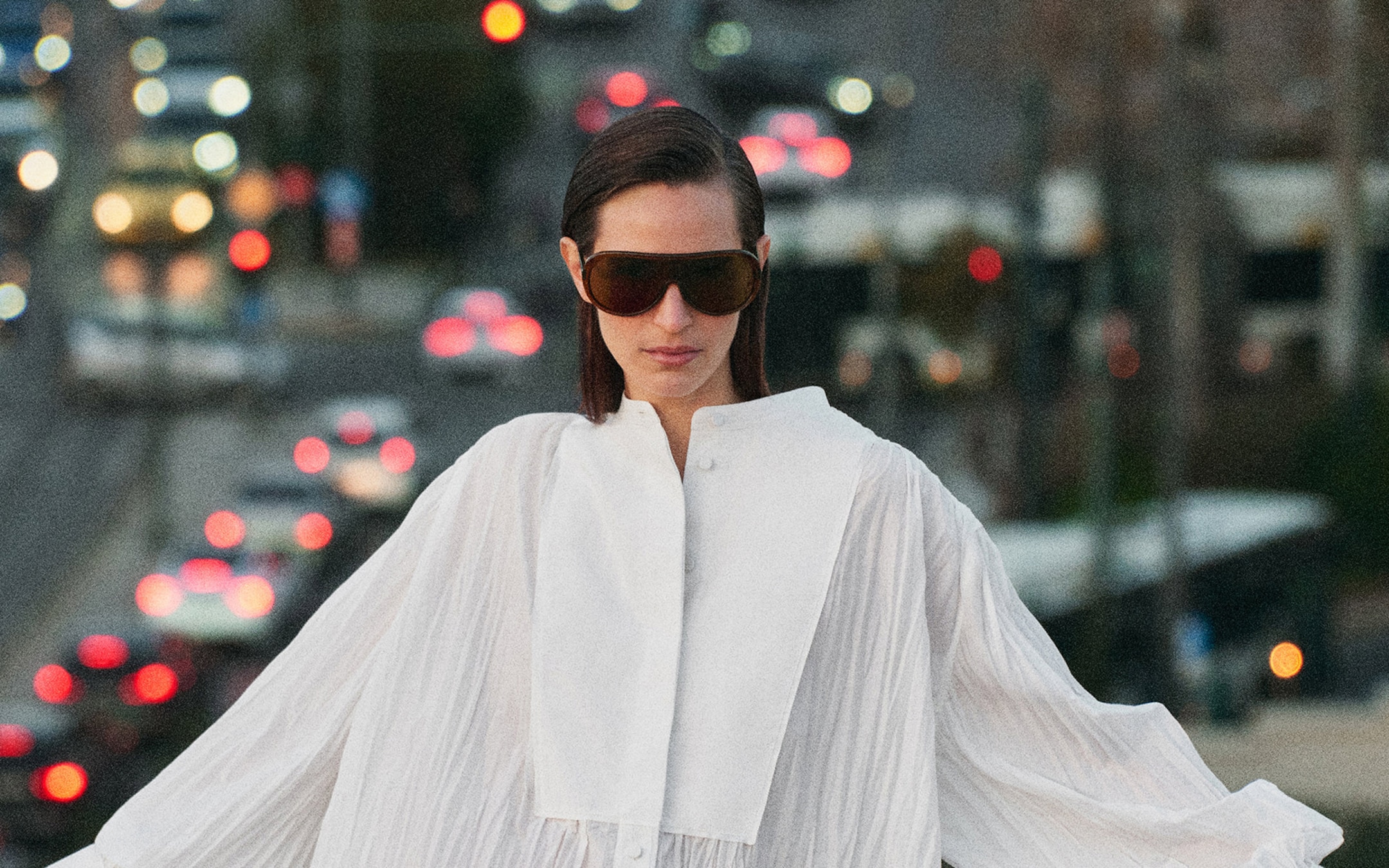 Person in a white flowing outfit and large sunglasses stands outdoors at dusk, blurred city lights in the background.